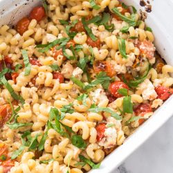 A white baking dish filled with baked pasta, cherry tomatoes, crumbled feta cheese, and fresh basil, set on a marble countertop.