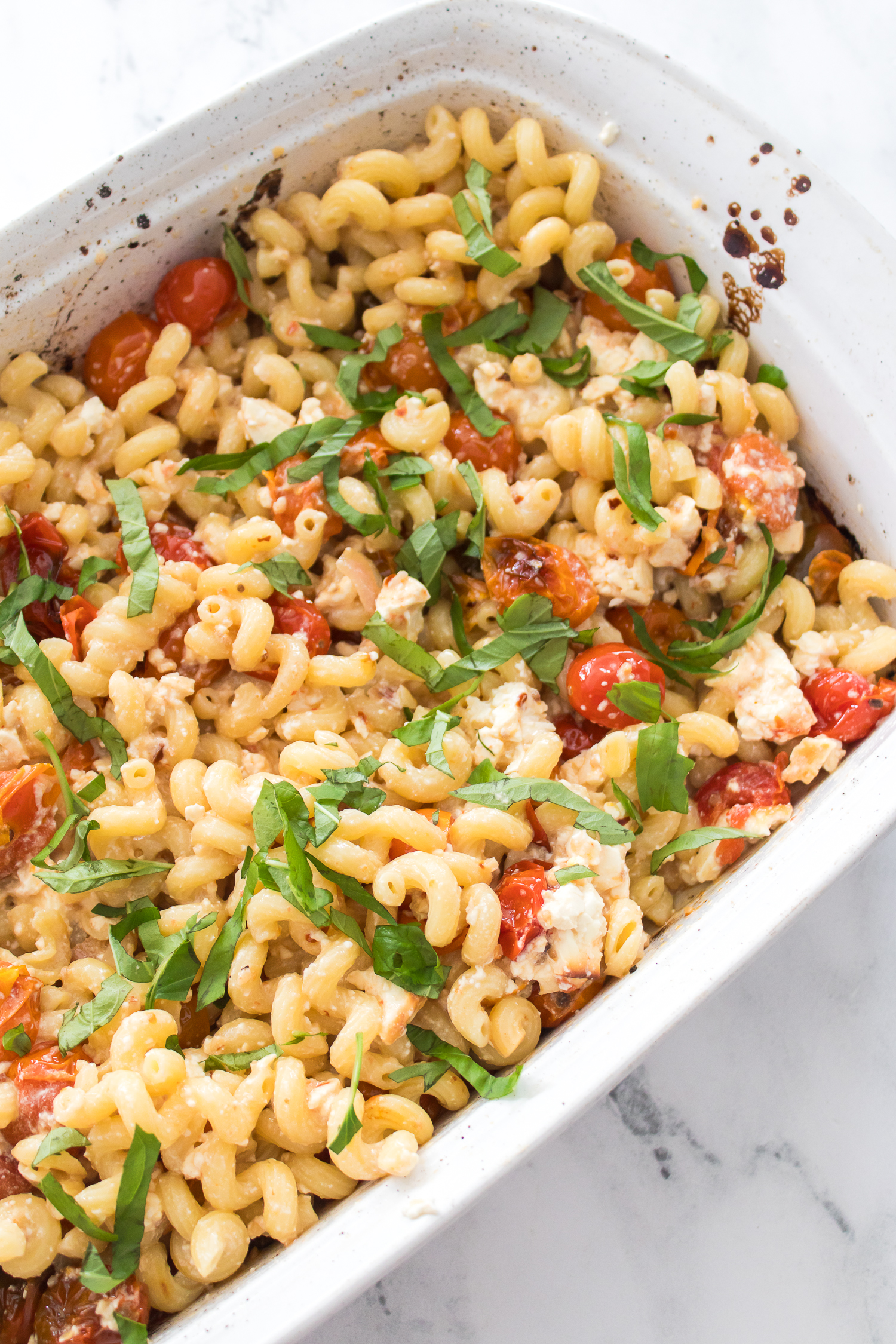 A white baking dish filled with baked pasta, cherry tomatoes, crumbled feta cheese, and fresh basil, set on a marble countertop.