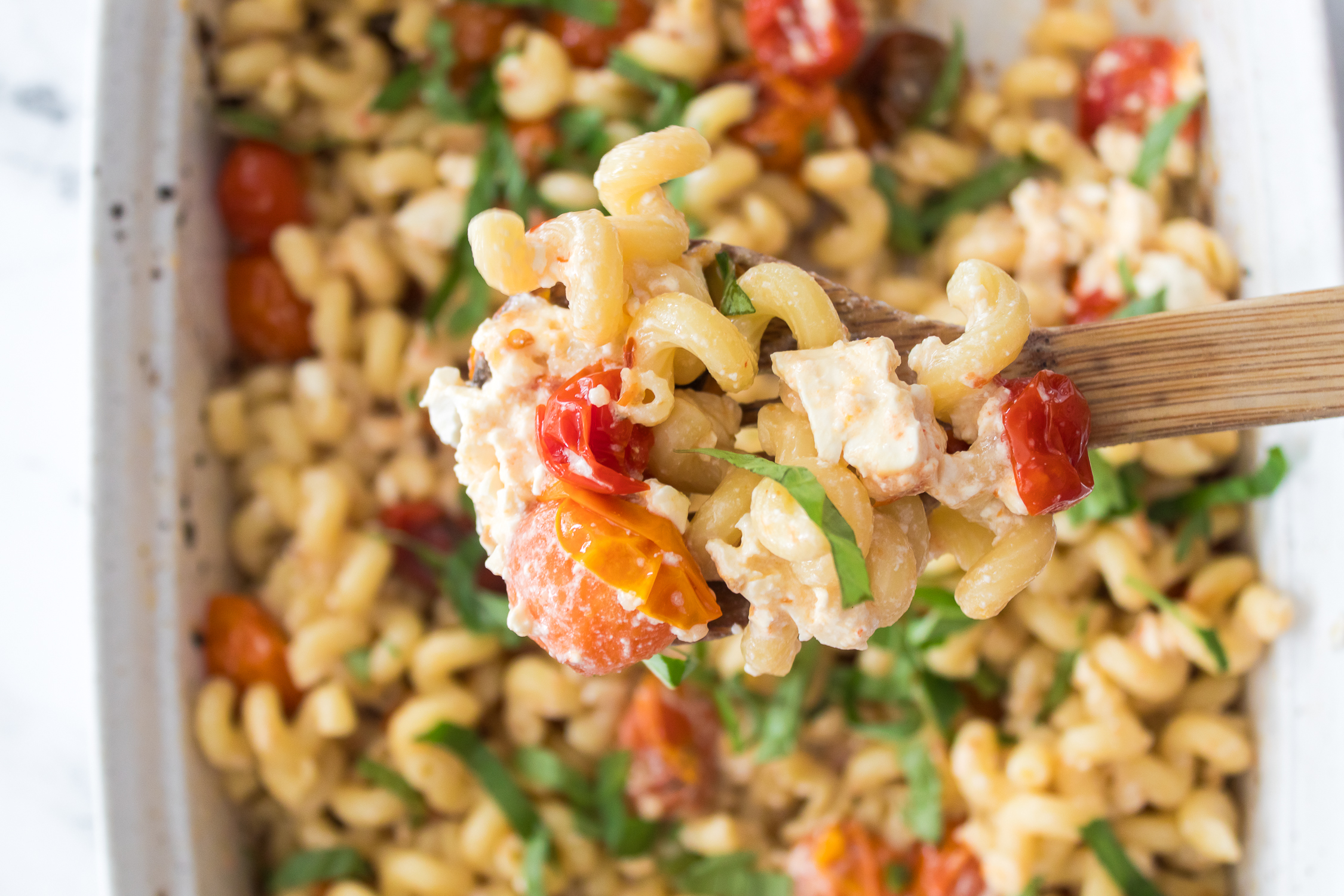 A wooden spoon holds a serving of baked pasta with fusilli, cherry tomatoes, crumbled feta cheese, and fresh basil over a casserole dish.