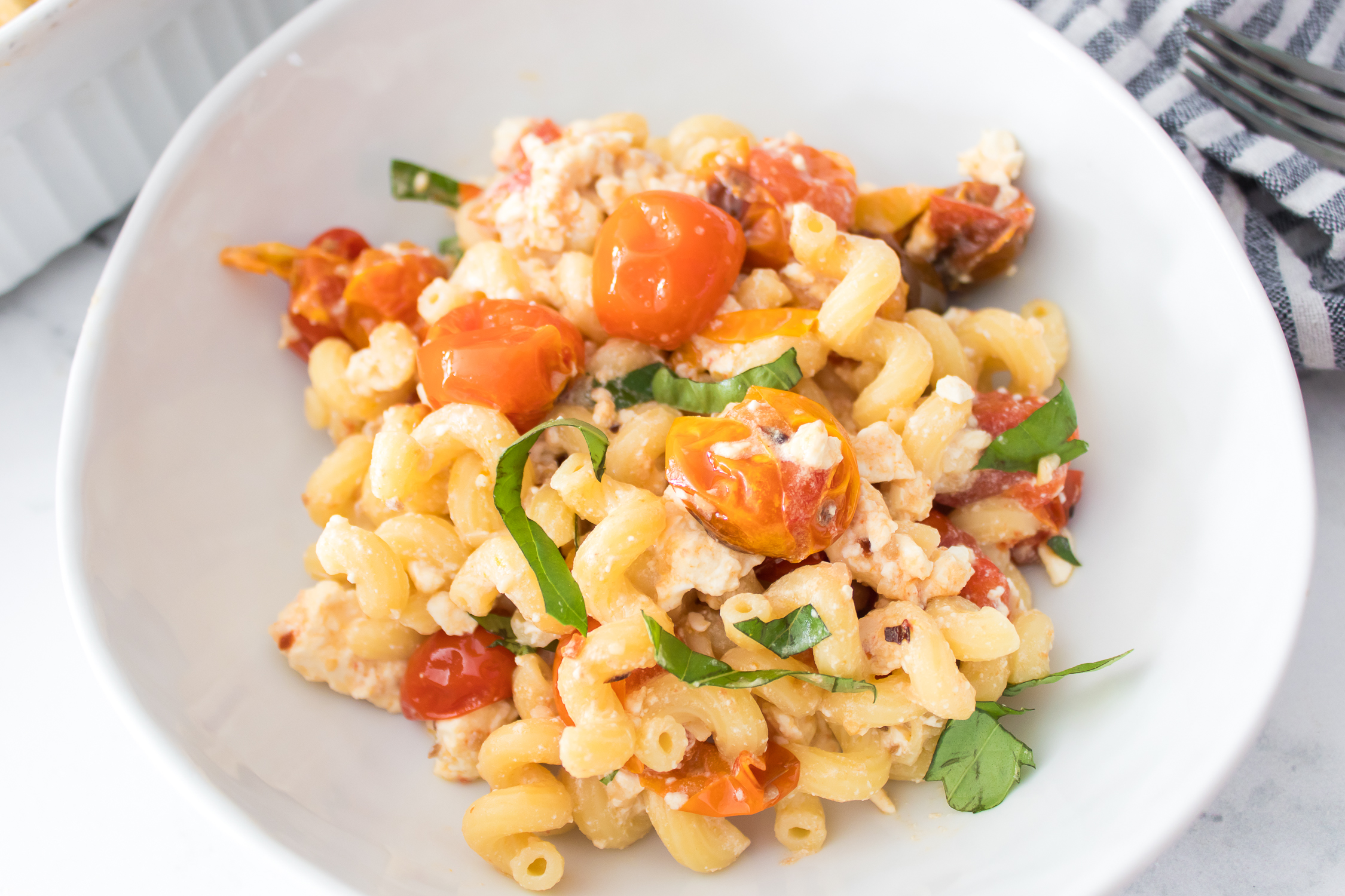A white bowl filled with cooked pasta, cherry tomatoes, crumbled feta cheese, and fresh basil leaves.
