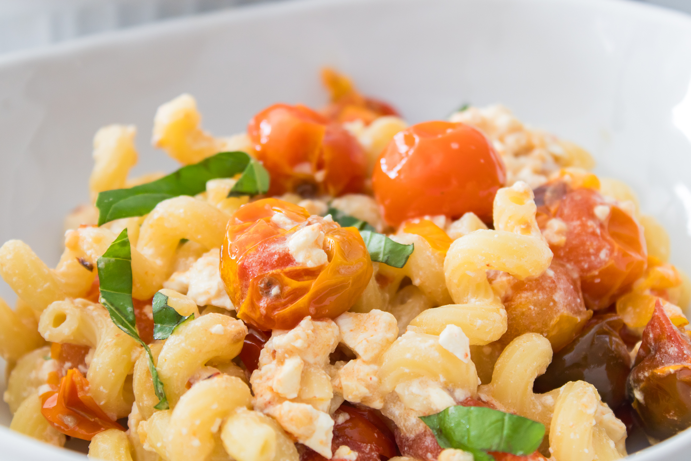 A close-up of pasta with roasted cherry tomatoes, crumbled feta cheese, and fresh basil in a white bowl.