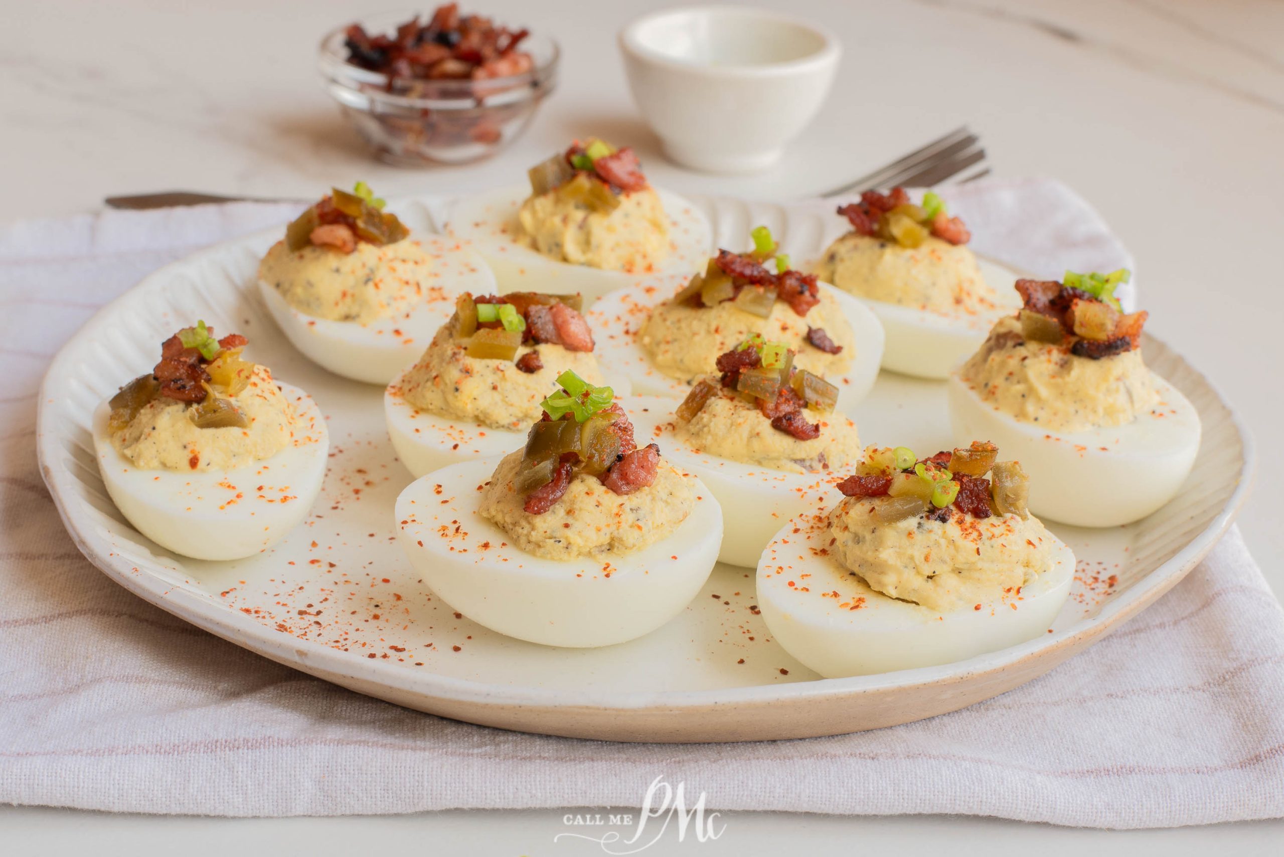 A plate of Jalapeno Popper Deviled Eggs topped with bacon bits and chopped green onions, set on a white napkin with a small bowl and fork in the background.