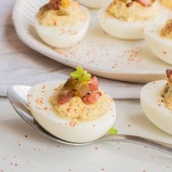 A spoon holds a Jalapeno Popper Deviled Egg topped with bacon and garnish. More deviled eggs are arranged on a white plate in the background.