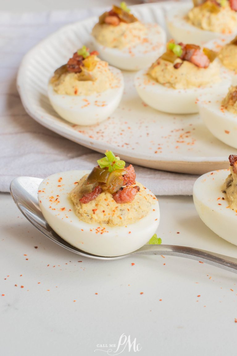 A spoon holds a Jalapeno Popper Deviled Egg topped with bacon and garnish. More deviled eggs are arranged on a white plate in the background.