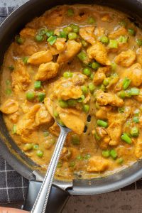 A pan filled with creamy chicken curry, green peas, and chopped green onions, with a spoon lifting a portion of the dish.