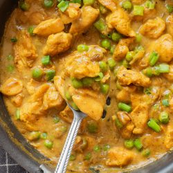 A pan filled with creamy chicken curry, green peas, and chopped green onions, with a spoon lifting a portion of the dish.