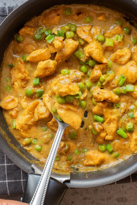 A pan filled with creamy chicken curry, green peas, and chopped green onions, with a spoon lifting a portion of the dish.