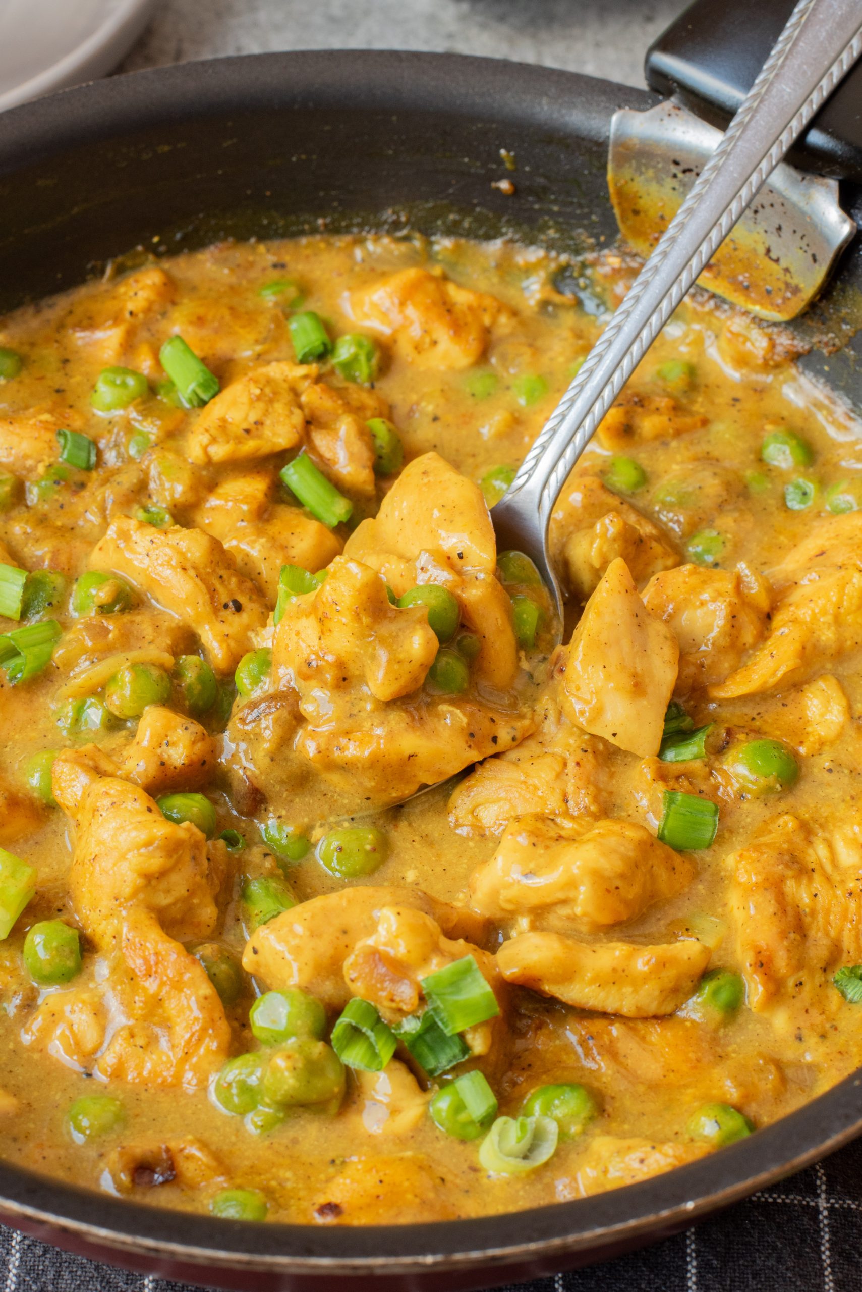 A pan filled with cooked chicken pieces in a yellow sauce, mixed with green peas and garnished with chopped green onions, with a spoon resting inside.