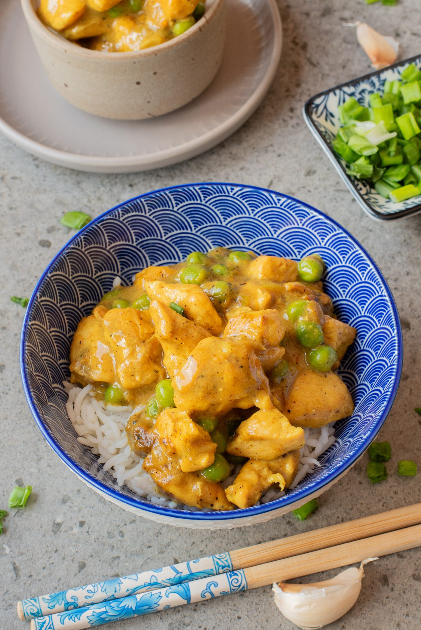 A blue patterned bowl filled with white rice, topped with yellow curry chicken and green peas, with chopsticks and chopped green onions nearby.