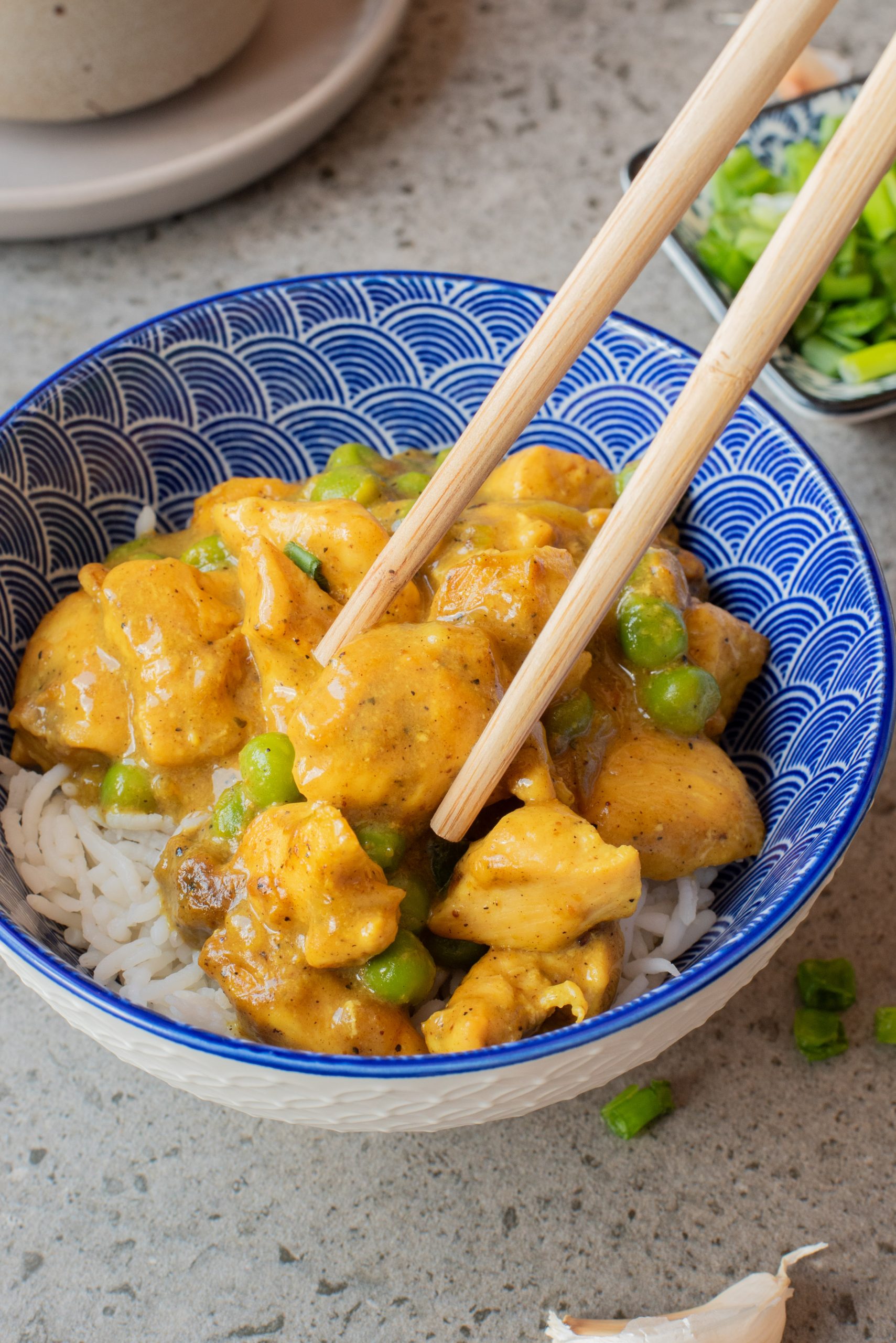 A blue-patterned bowl of white rice topped with yellow curry chicken and green peas, with chopsticks picking up a piece.