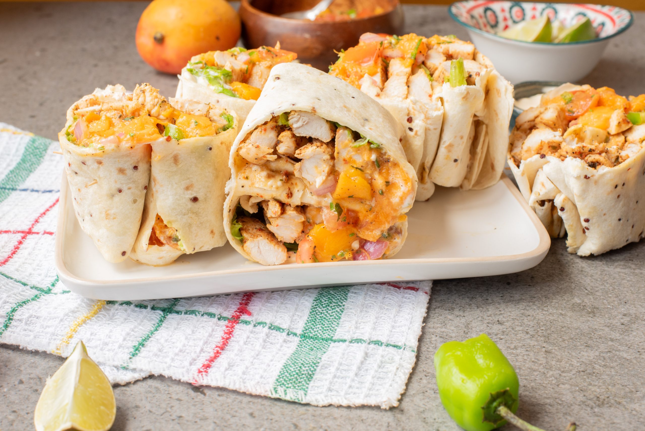 Four Mango Salsa Jerk Chicken Wraps filled with grilled chicken, diced mango, and vegetables are arranged on a rectangular white plate, with a lime wedge and green chili pepper nearby.