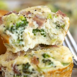 Two Muffin Tin Egg Bites with broccoli and ham are stacked, the top one partially bitten to reveal the fluffy interior.
