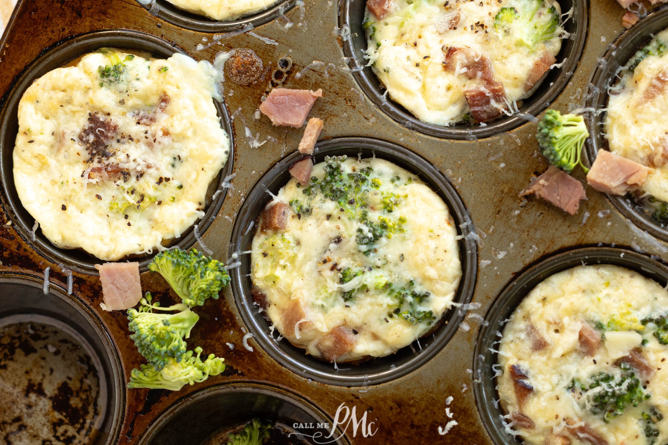 Muffin tin egg bites filled with broccoli, ham, and cheese. Shredded cheese and broccoli florets are scattered on and around the muffins for a tasty, colorful touch.