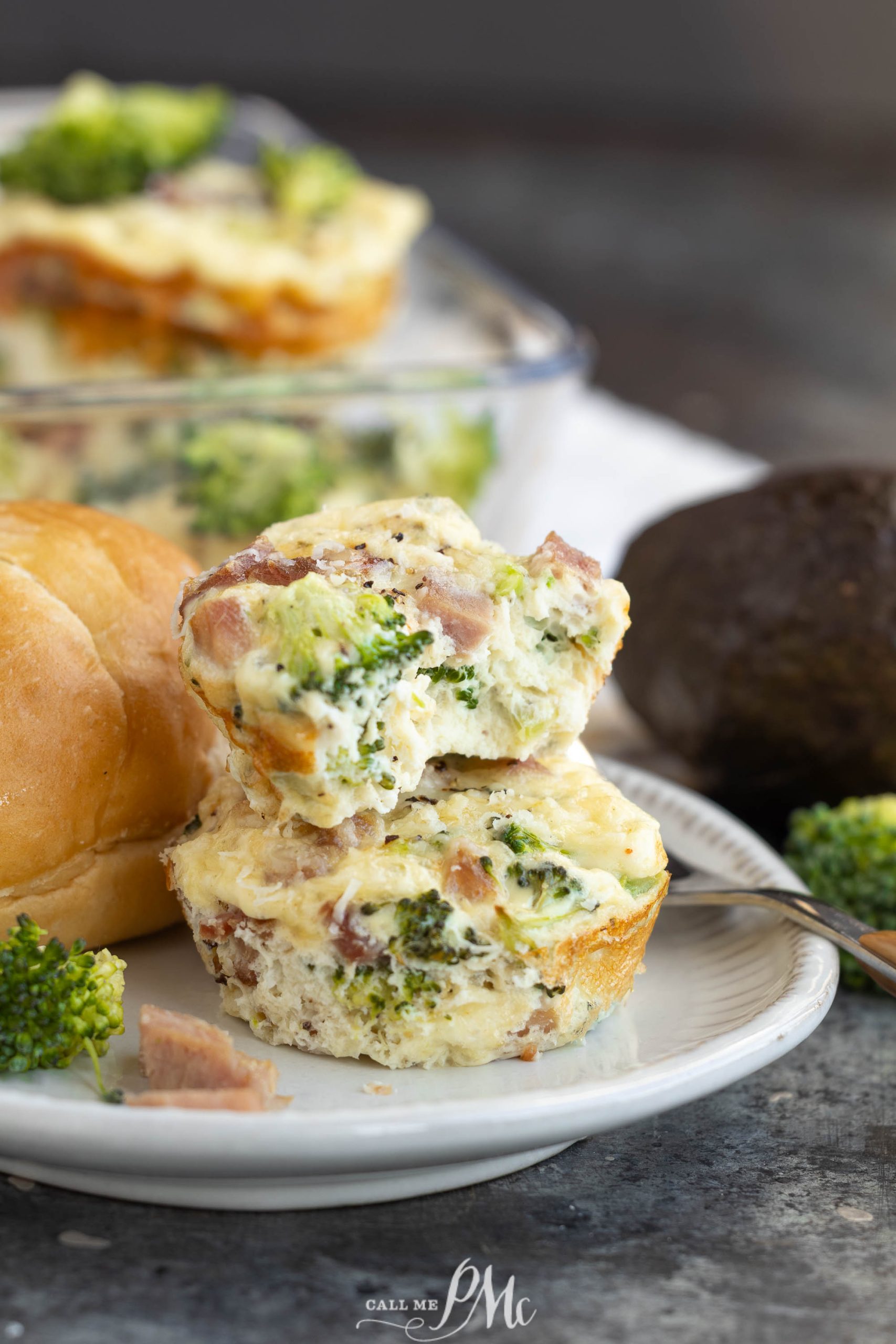 Two broccoli and ham Muffin Tin Egg Bites stacked on a plate with a roll, fresh broccoli, and part of an avocado in the background.