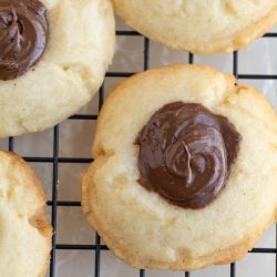 Three Nutella shortbread thumbprint cookies with a dollop of melted chocolate in the center are cooling on a black wire rack.