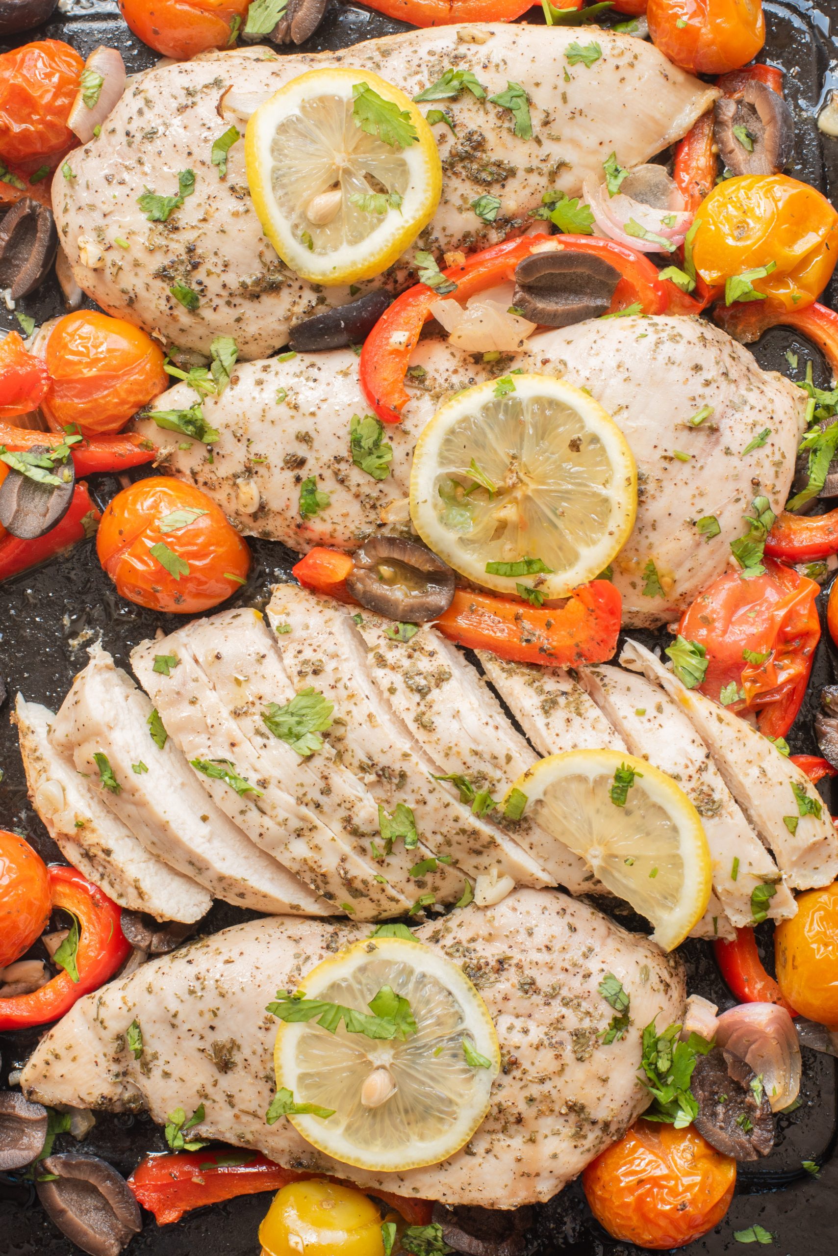 Baked chicken breasts with lemon slices, cherry tomatoes, red bell peppers, mushrooms, and chopped herbs on a baking tray.
