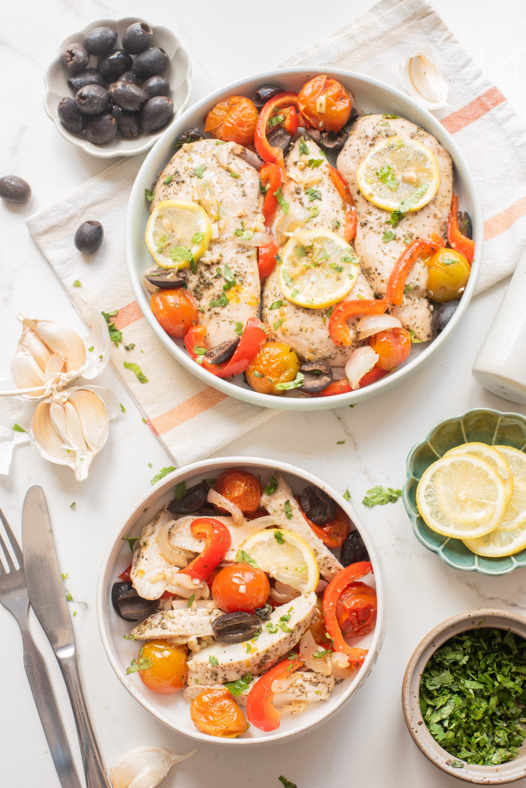 Two bowls of baked chicken breasts with lemon slices, cherry tomatoes, red bell peppers, onions, and black olives, garnished with herbs. Sliced garlic, lemon, and fresh herbs on the side.