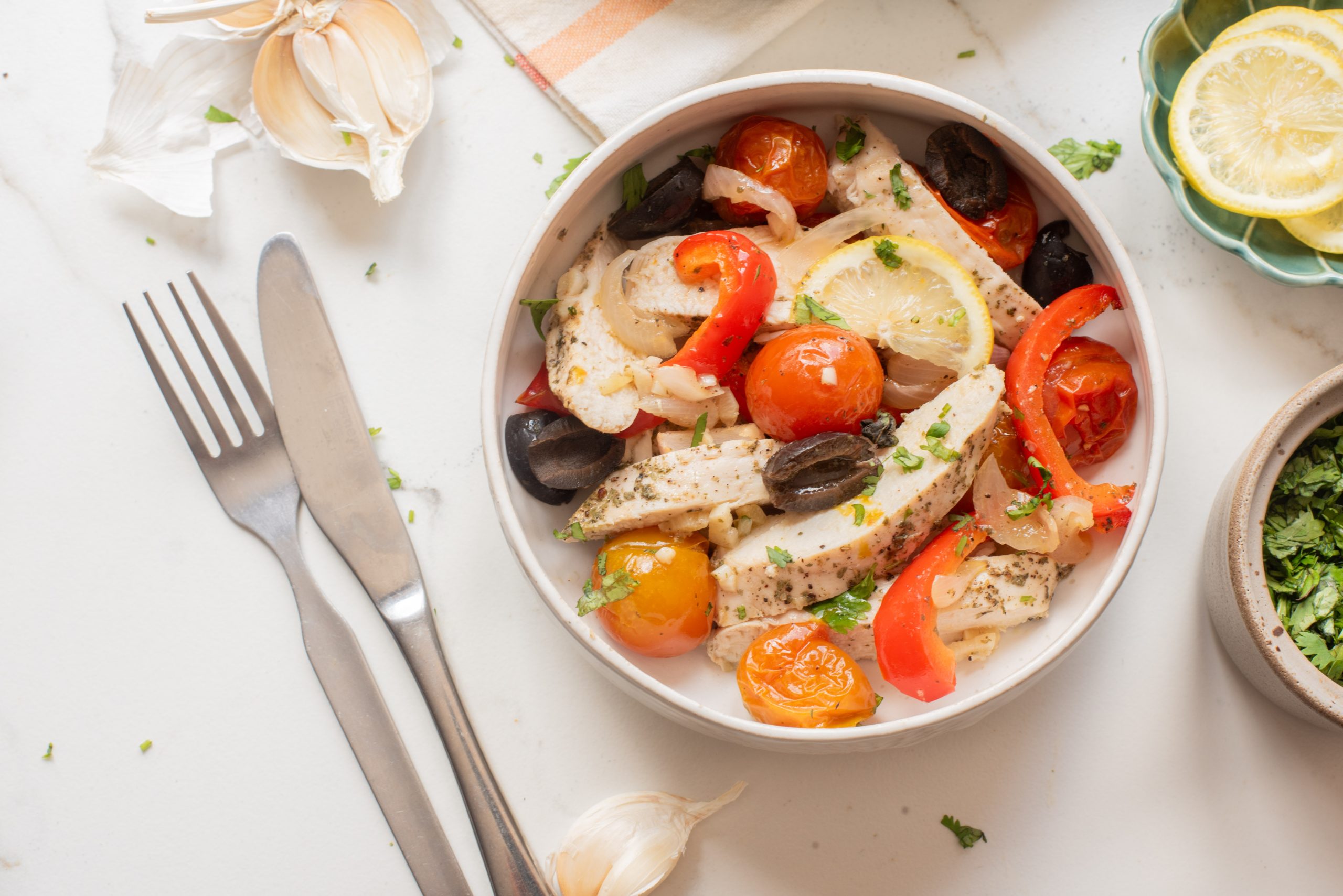 A bowl of cooked chicken breast with cherry tomatoes, black olives, red bell peppers, lemon slices, and herbs on a white table next to a fork, knife, garlic, and lemon slices.