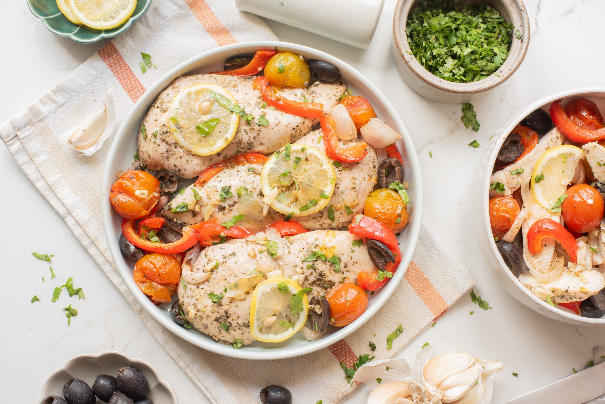 A plate of Sheet Pan Mediterranean Chicken Breasts topped with lemon slices, cherry tomatoes, red bell peppers, olives, onions, and chopped herbs.