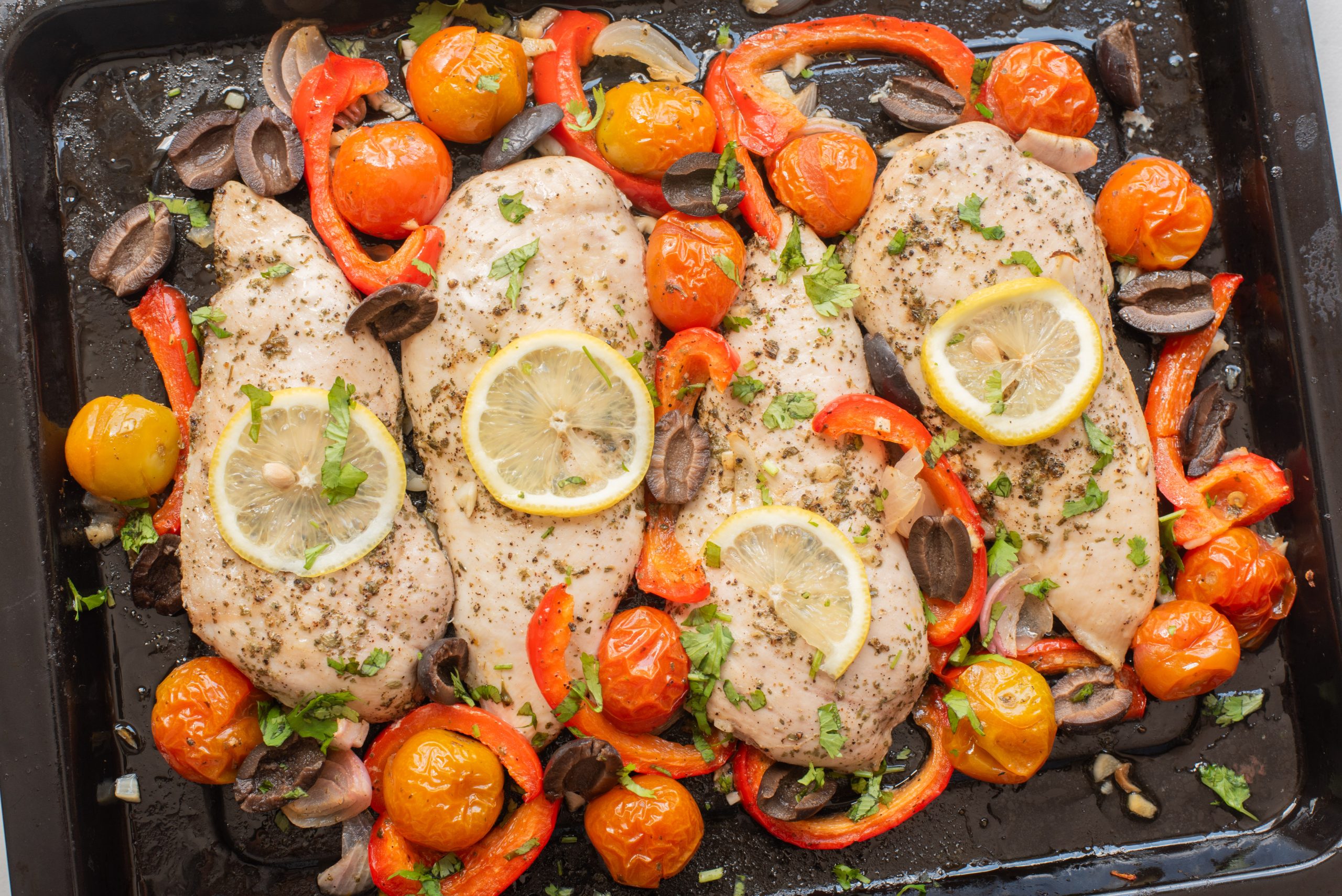 Four seasoned chicken breasts topped with lemon slices are baked on a tray with cherry tomatoes, sliced red bell peppers, mushrooms, and herbs.