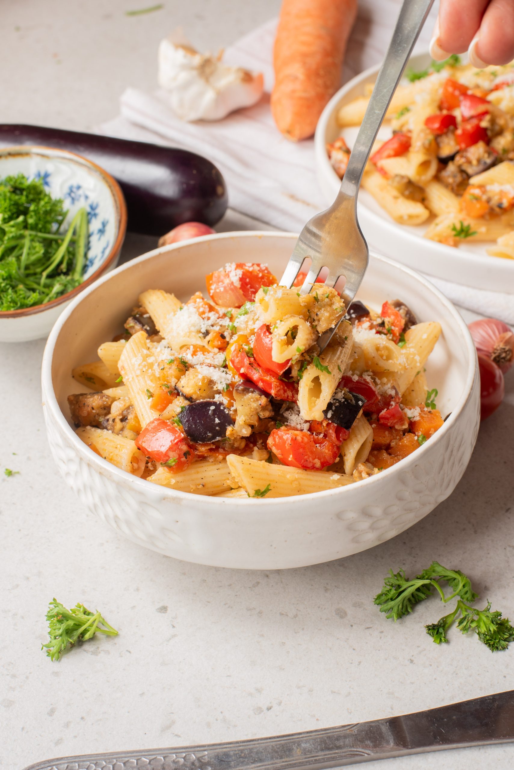 A hand uses a fork to pick up Sheet Pan Mediterranean Vegetable Pasta from a white bowl on a light surface, surrounded by fresh vegetables and herbs.