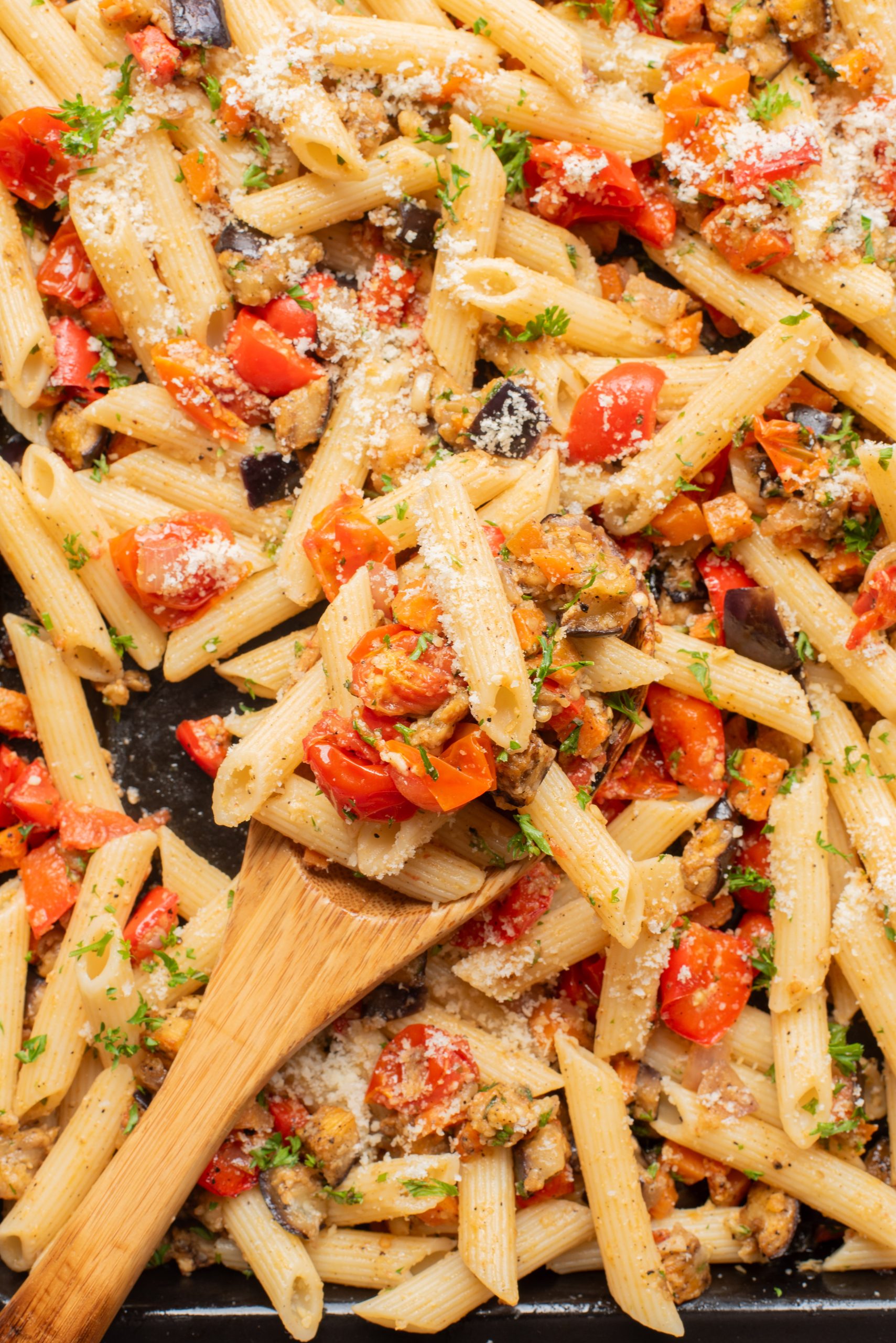 Baked penne pasta with diced tomatoes, eggplant, zucchini, parsley, and grated cheese, served on a wooden spoon over a sheet pan.