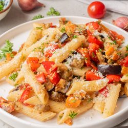 A plate of penne pasta mixed with diced eggplant, tomatoes, red peppers, and herbs, topped with grated cheese.