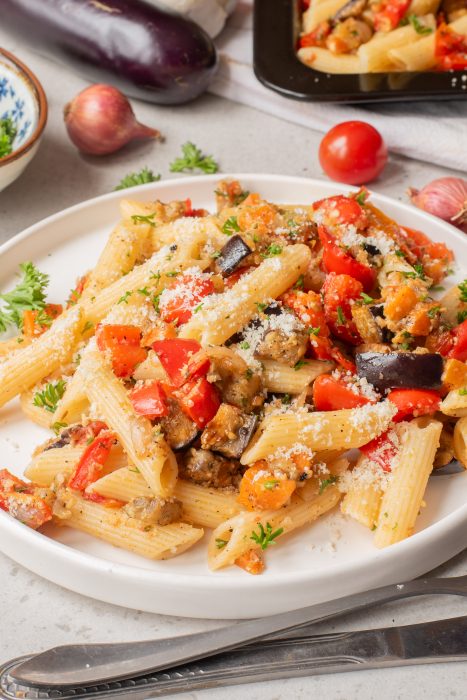 A plate of penne pasta mixed with diced eggplant, tomatoes, red peppers, and herbs, topped with grated cheese.