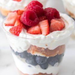 A clear plastic cup filled with layers of whipped cream, blueberries, cake, and topped with strawberries and raspberries.