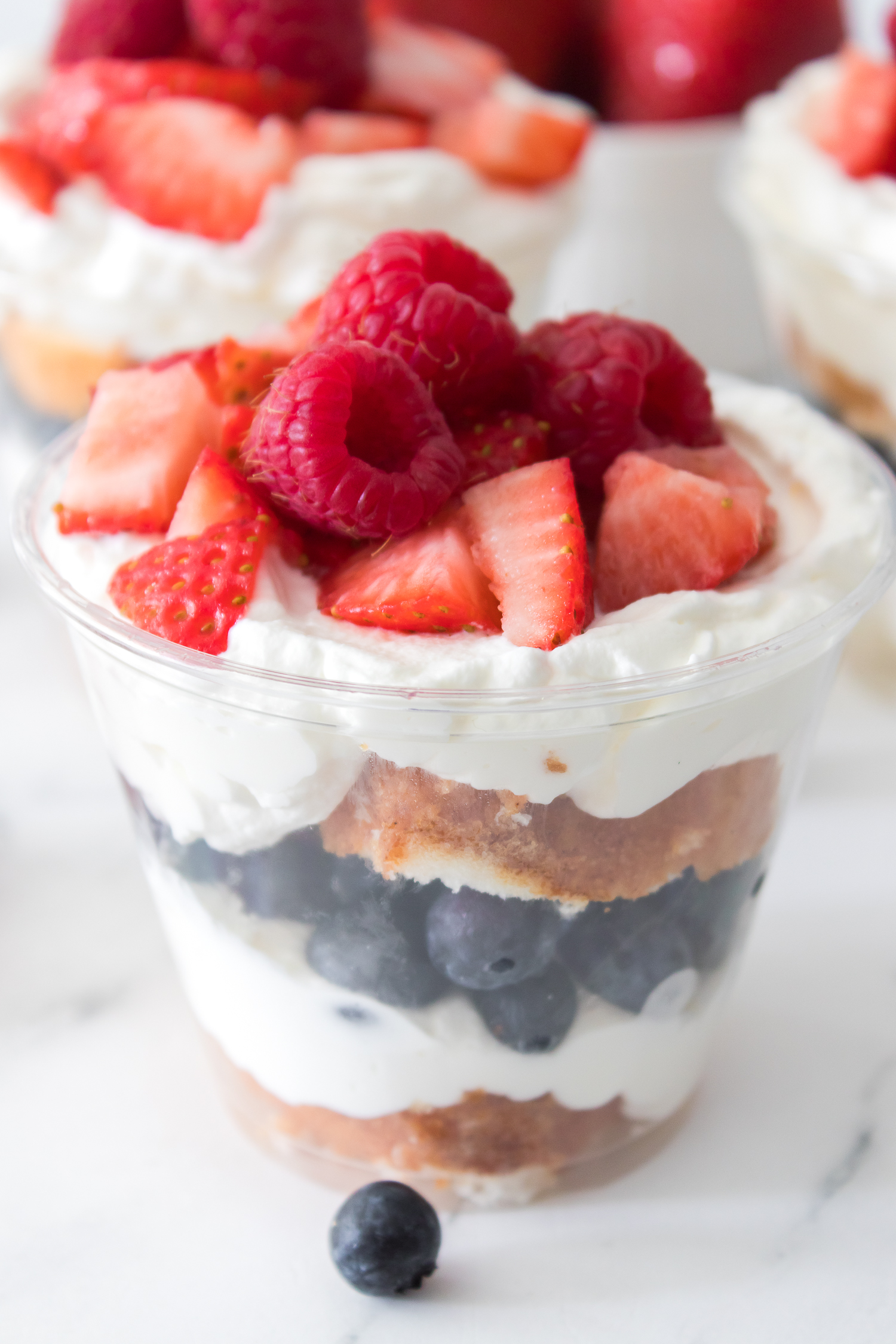 A clear plastic cup filled with layers of whipped cream, blueberries, cake, and topped with strawberries and raspberries.