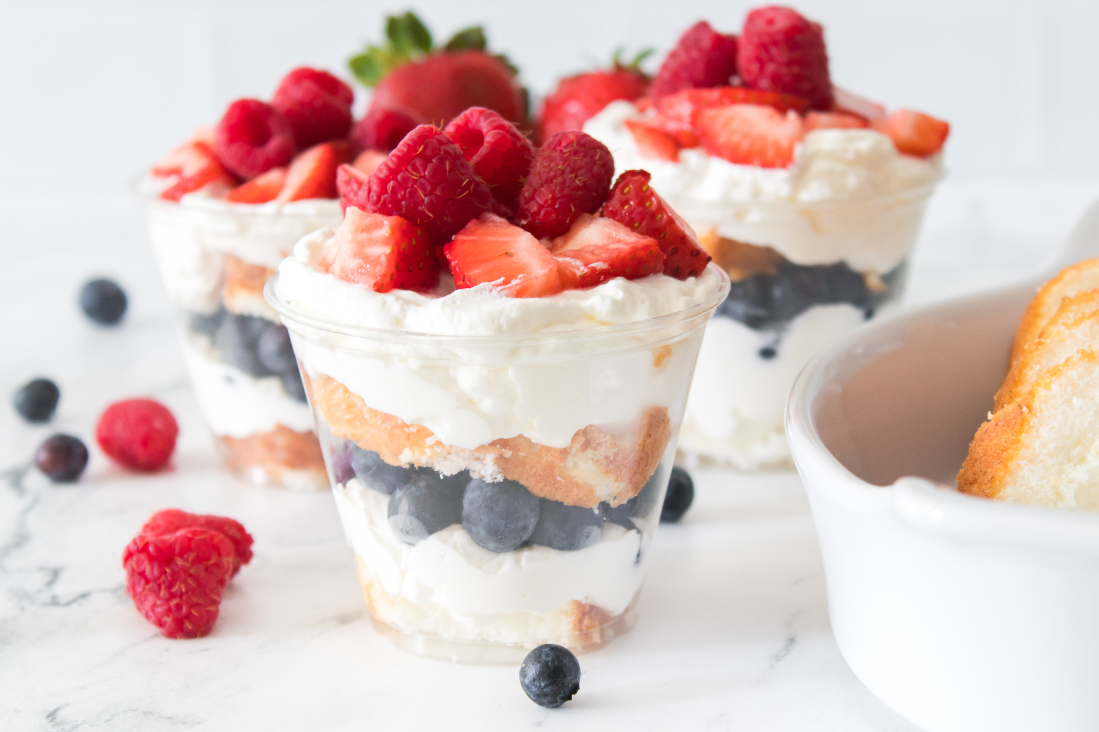 Three clear cups layered with whipped cream, blueberries, cake pieces, raspberries, and strawberries are arranged on a white surface with loose berries and a cake dish nearby.