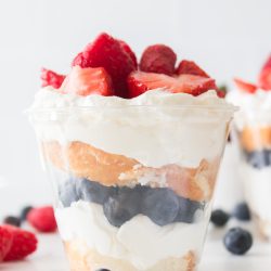 A clear cup filled with layers of whipped cream, blueberries, cake, and topped with sliced strawberries and raspberries.
