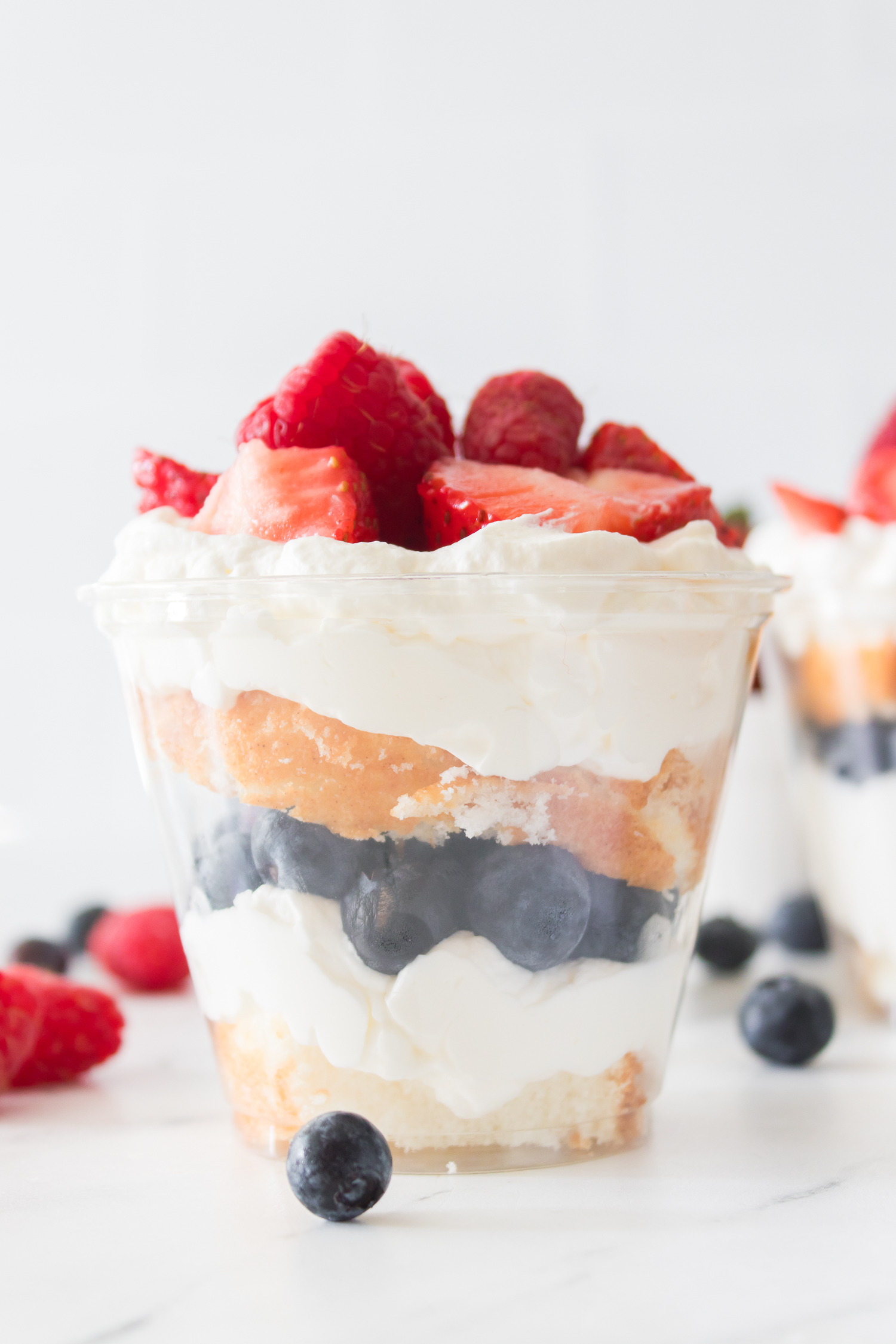 A clear cup filled with layers of whipped cream, blueberries, cake, and topped with sliced strawberries and raspberries.