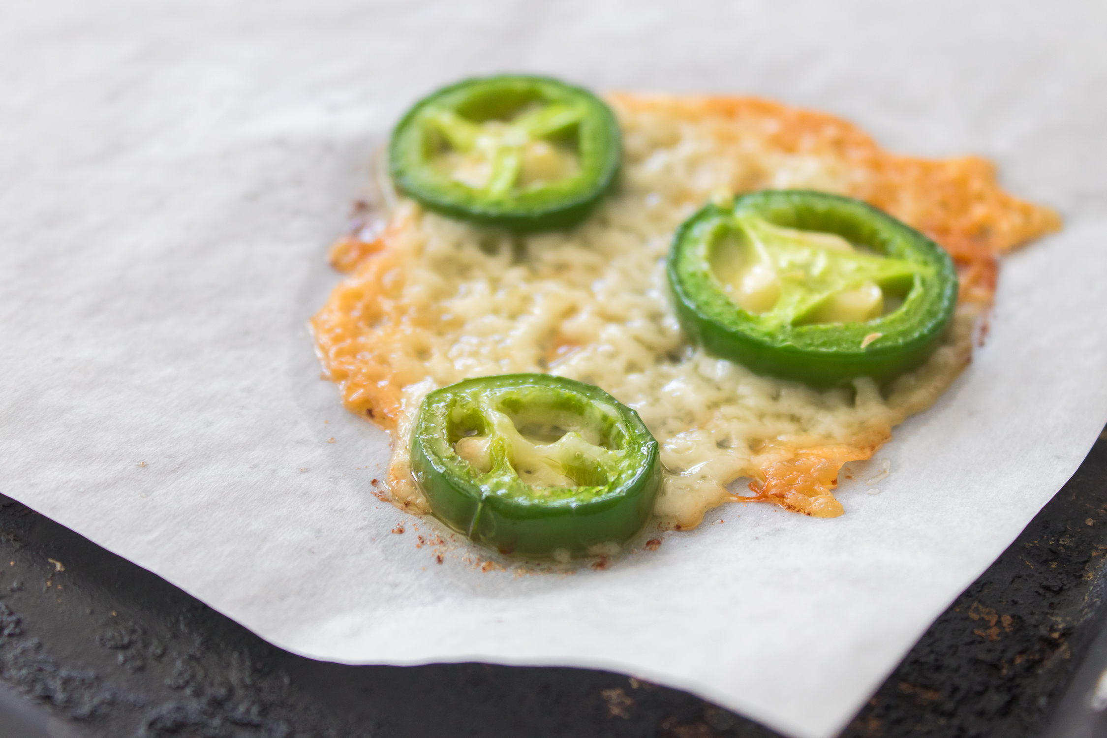 A piece of melted cheese topped with four slices of fresh jalapeño pepper rests on a sheet of parchment paper.