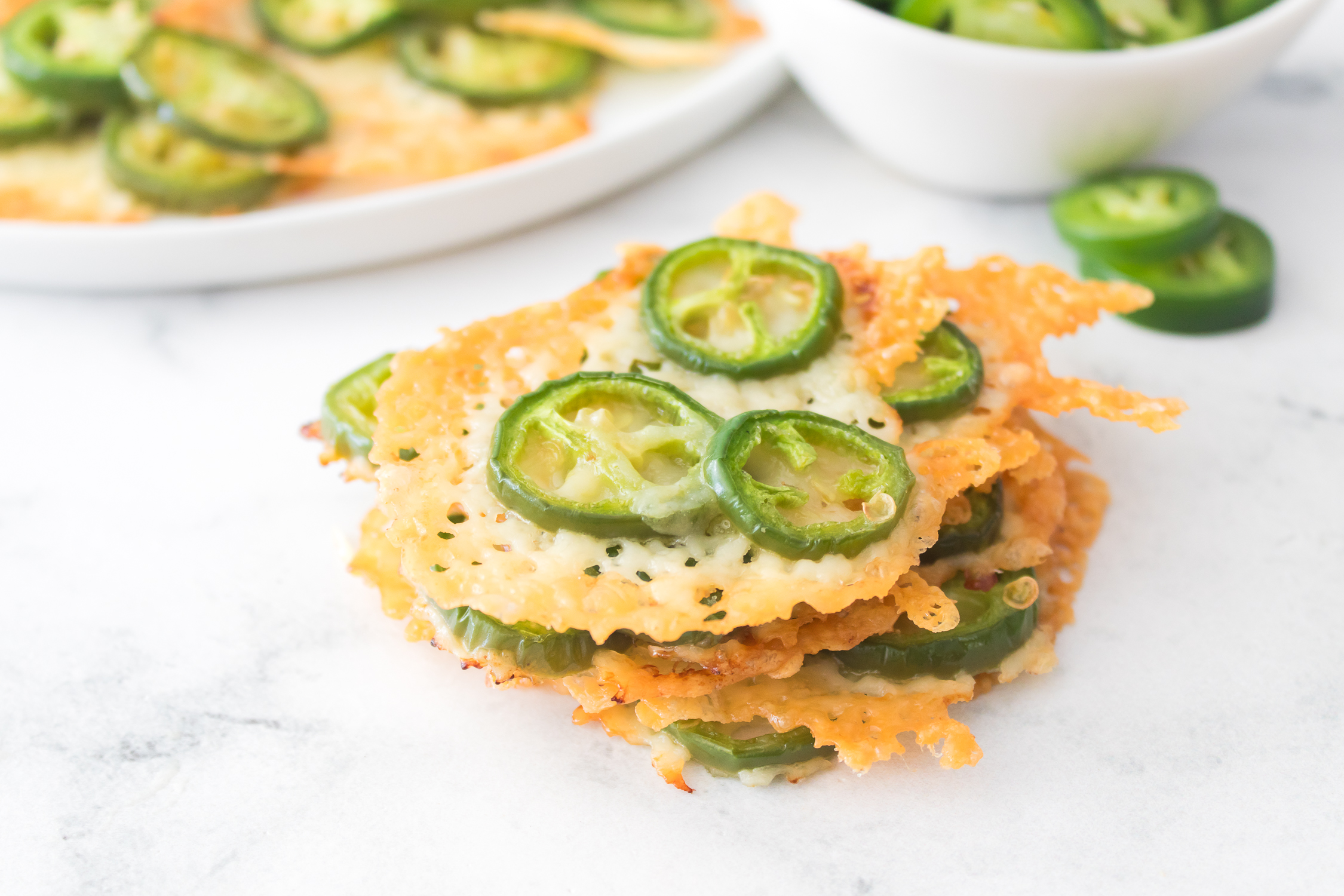 A stack of Keto Jalapeno Cheese Crisps topped with sliced jalapeños, with more crisps and a bowl of jalapeños in the background.