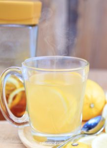 A clear glass mug filled with hot lemon tea, reminiscent of Starbucks Medicine Ball Tea, sits on a table with fresh lemons, a spoon, and a jar of honey in the background. Steam gently rises from the mug.