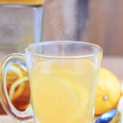 A clear glass mug filled with hot lemon tea, reminiscent of Starbucks Medicine Ball Tea, sits on a table with fresh lemons, a spoon, and a jar of honey in the background. Steam gently rises from the mug.