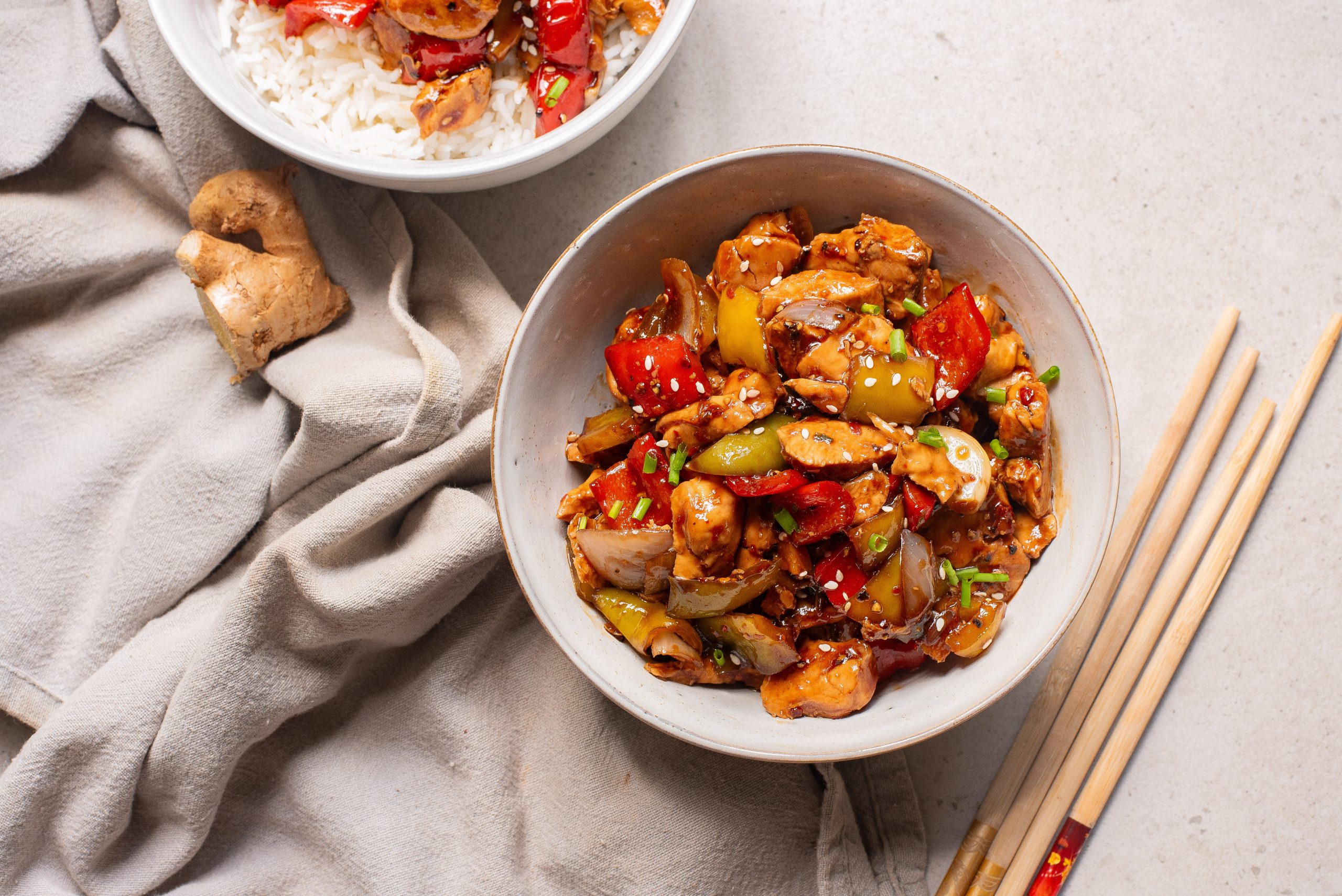 A delicious Black Pepper Chicken Bowl featuring stir-fried chicken with bell peppers and onions in sauce, served beside a bowl of rice, chopsticks, a beige cloth, and fresh ginger on a light surface.