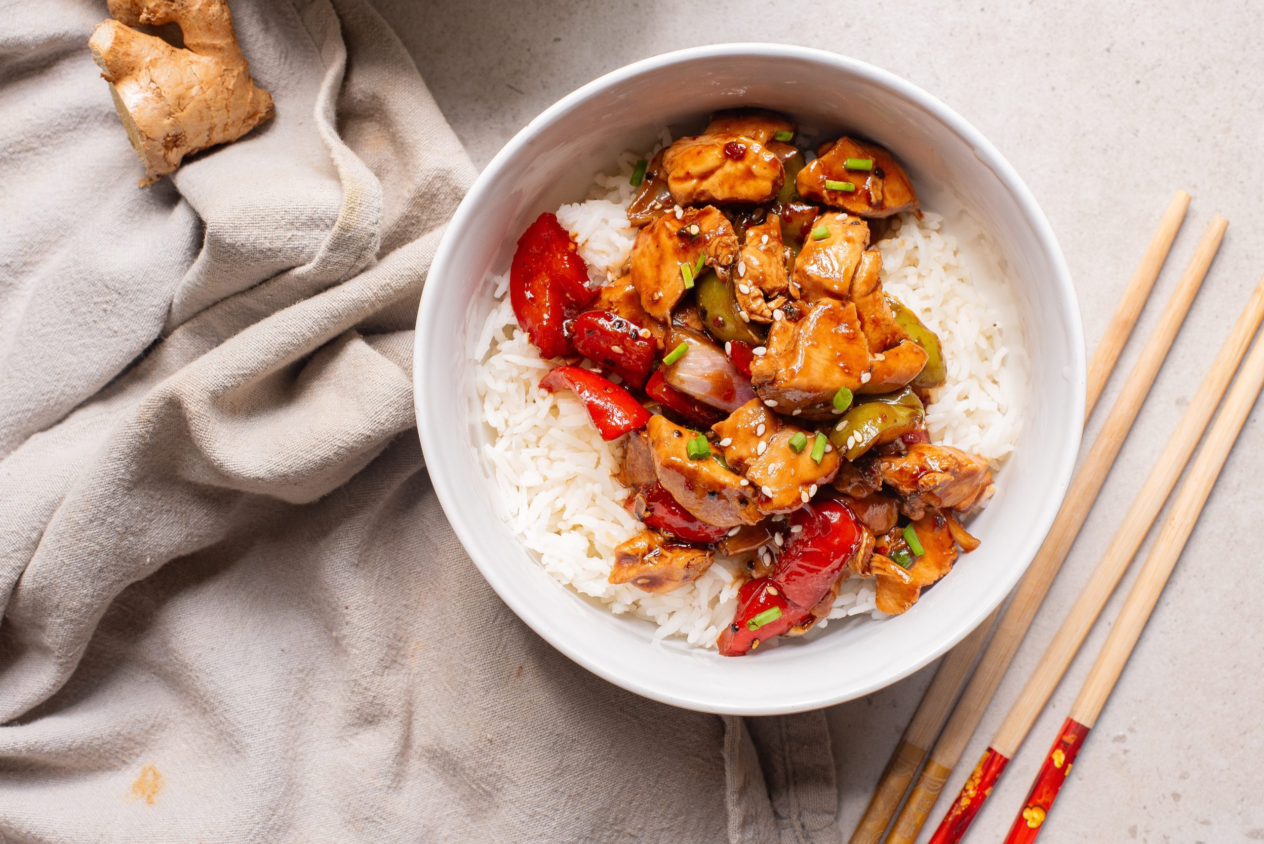 A bowl of white rice topped with Black Pepper Chicken Bowl stir-fried chicken, red bell peppers, onions, and sesame seeds, with chopsticks and a beige cloth napkin nearby.