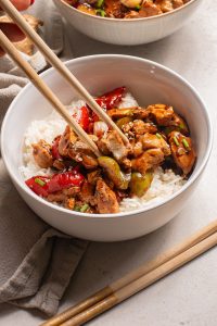A Black Pepper Chicken Bowl featuring white rice topped with stir-fried chicken, red bell peppers, and green vegetables, with chopsticks picking up a flavorful bite.