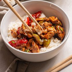 A Black Pepper Chicken Bowl featuring white rice topped with stir-fried chicken, red bell peppers, and green vegetables, with chopsticks picking up a flavorful bite.