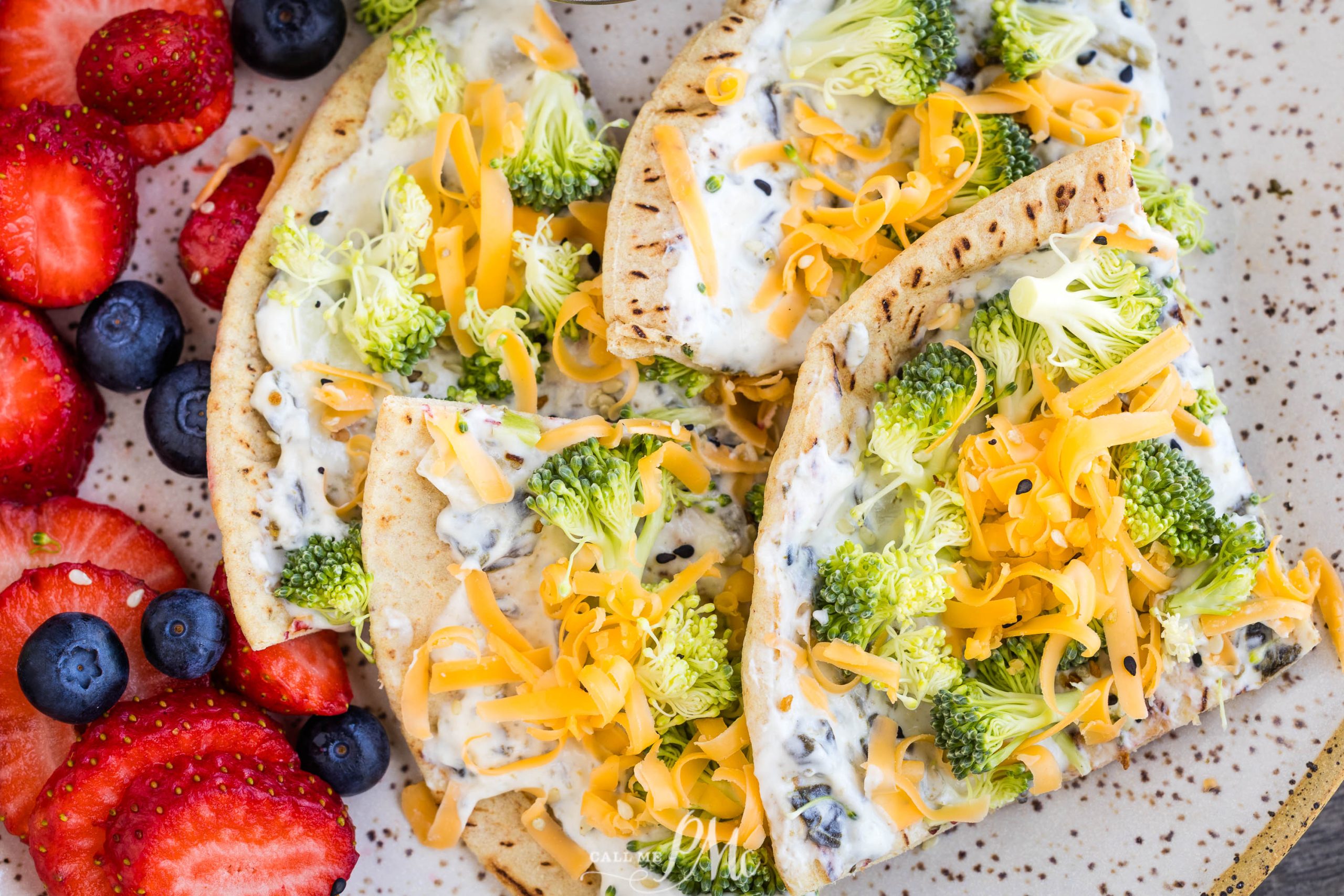 Pita bread slices topped with cream cheese, chopped broccoli, and shredded cheddar cheese, served with fresh strawberries and blueberries on a plate.