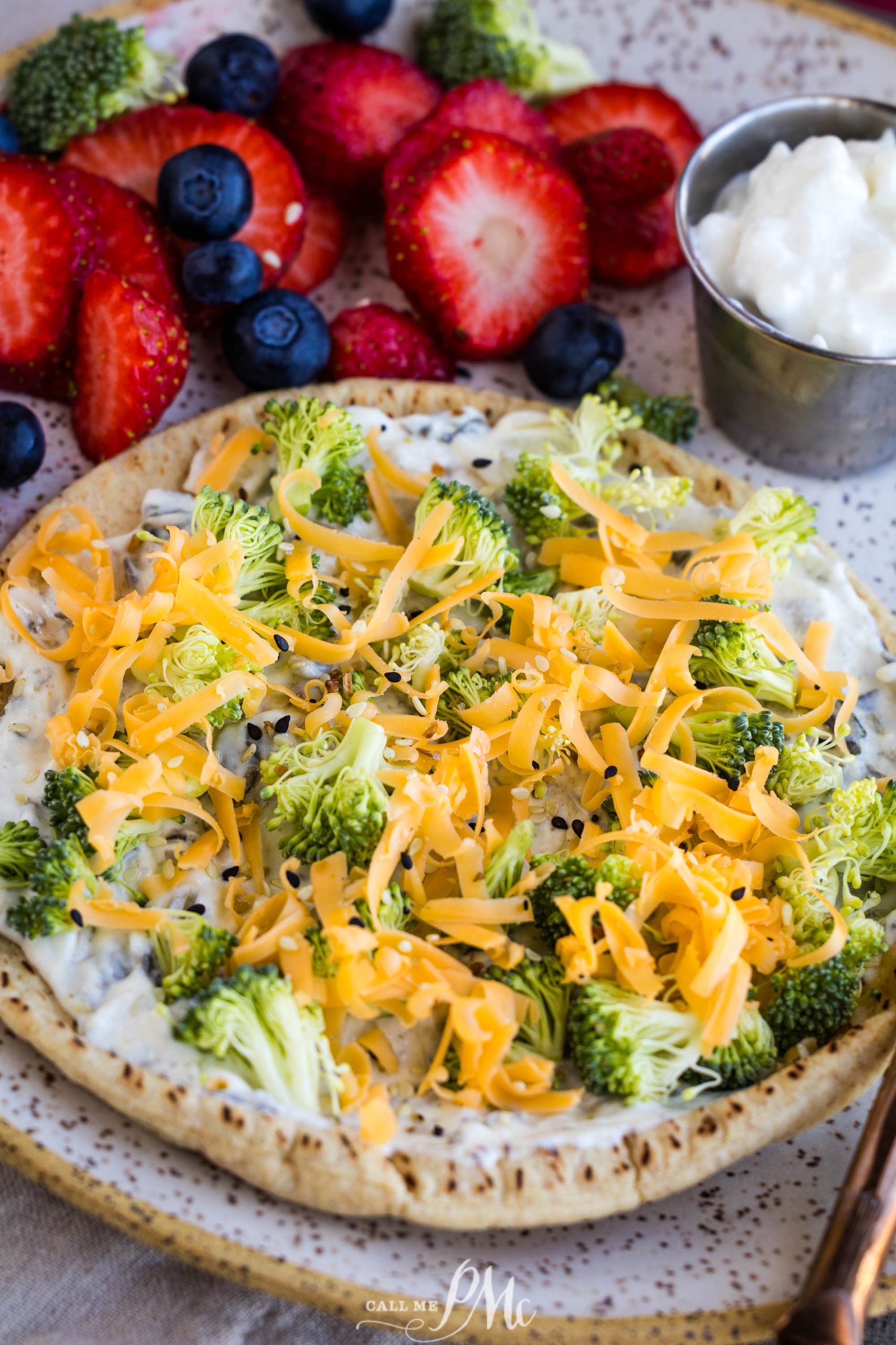 A plate with a veggie flatbread topped with broccoli, shredded cheddar cheese, and creamy spread, served with fresh strawberries, blueberries, and a small cup of dip.