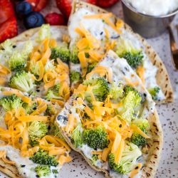 Flatbread topped with cream cheese, raw broccoli florets, and shredded cheddar cheese, served with a side of strawberries, blueberries, and a small cup of white dip.