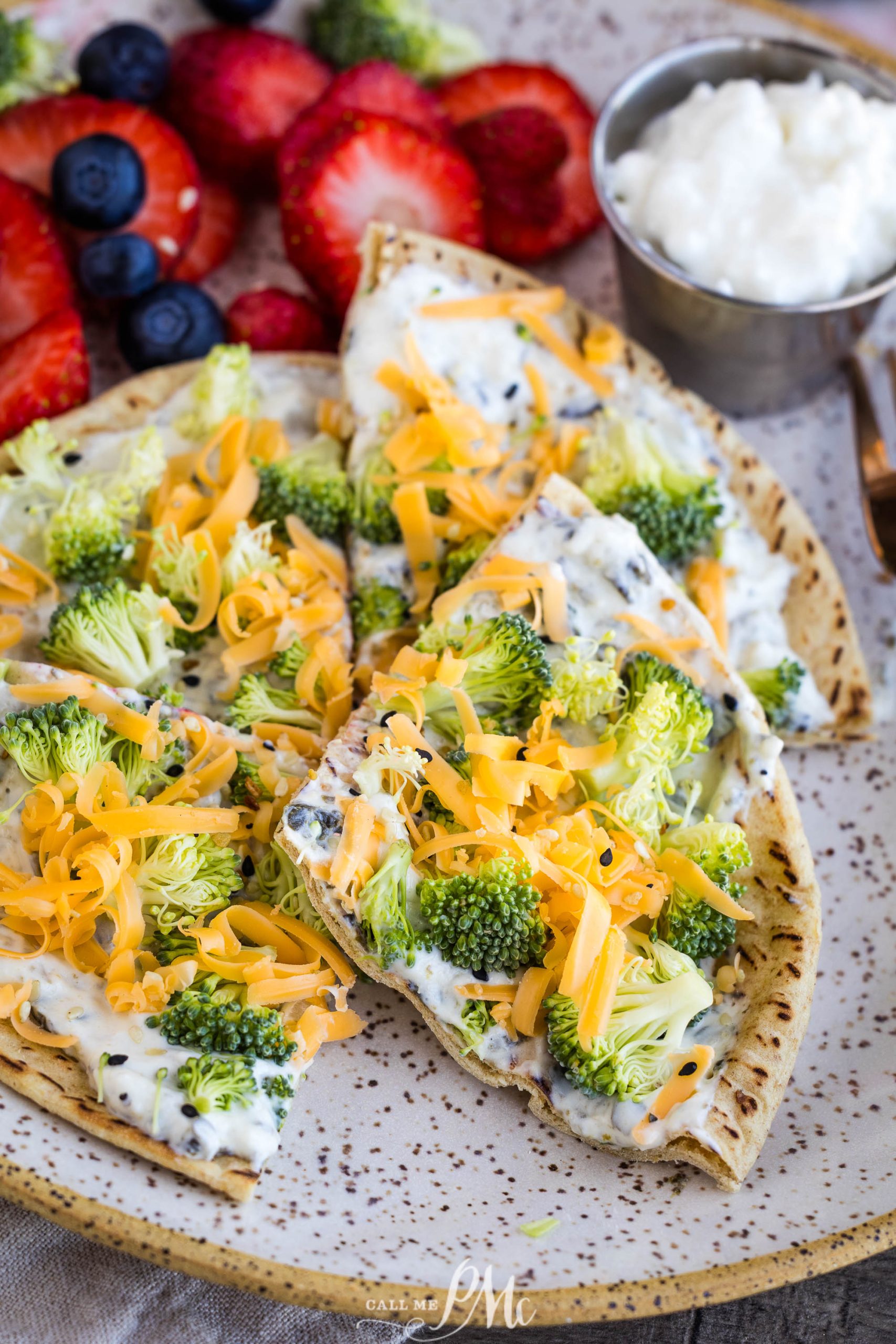 Flatbread topped with cream cheese, raw broccoli florets, and shredded cheddar cheese, served with a side of strawberries, blueberries, and a small cup of white dip.
