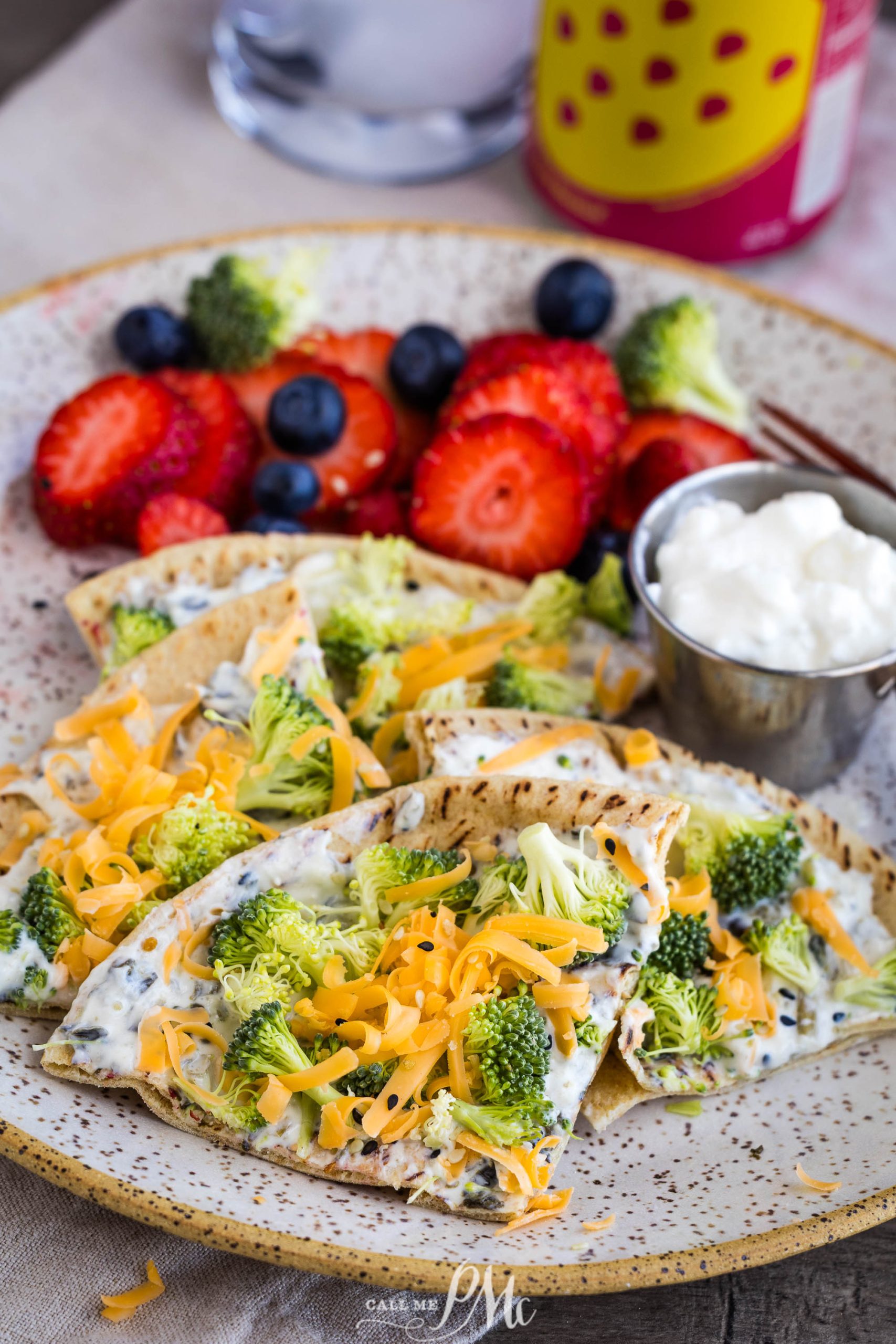 A plate with flatbread topped with broccoli, shredded cheese, and herbs, served with fresh strawberries, blueberries, and a side of cottage cheese.