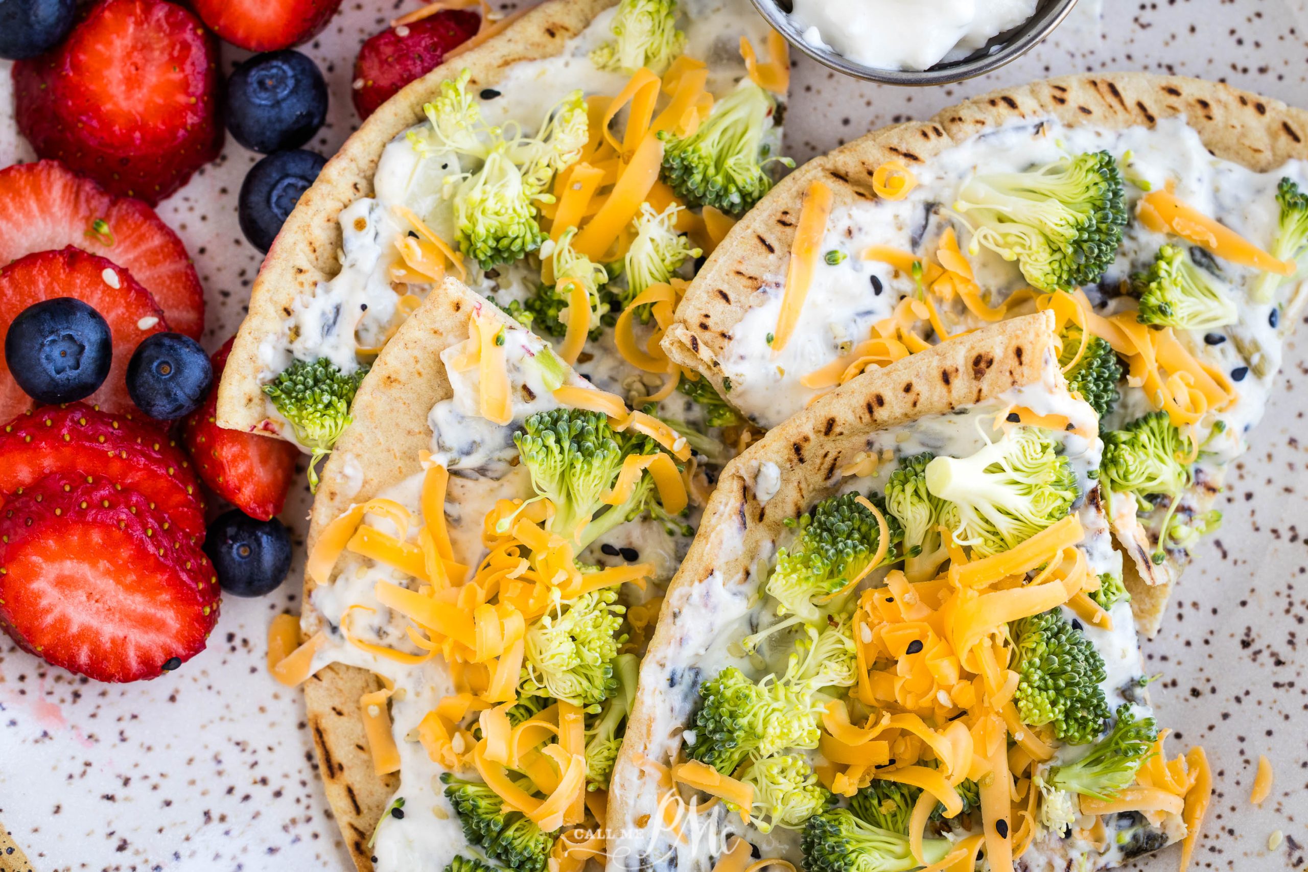 A plate with pita bread topped with cream cheese, raw broccoli, and shredded cheddar cheese, served with a side of fresh strawberries and blueberries.