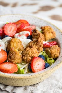 A bowl of Chicken Berry Poppy Seed Salad topped with fried chicken pieces, sliced strawberries, blueberries, shredded cheese, and a drizzle of creamy dressing.