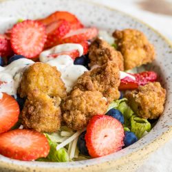 A bowl of Chicken Berry Poppy Seed Salad topped with fried chicken pieces, sliced strawberries, blueberries, shredded cheese, and a drizzle of creamy dressing.