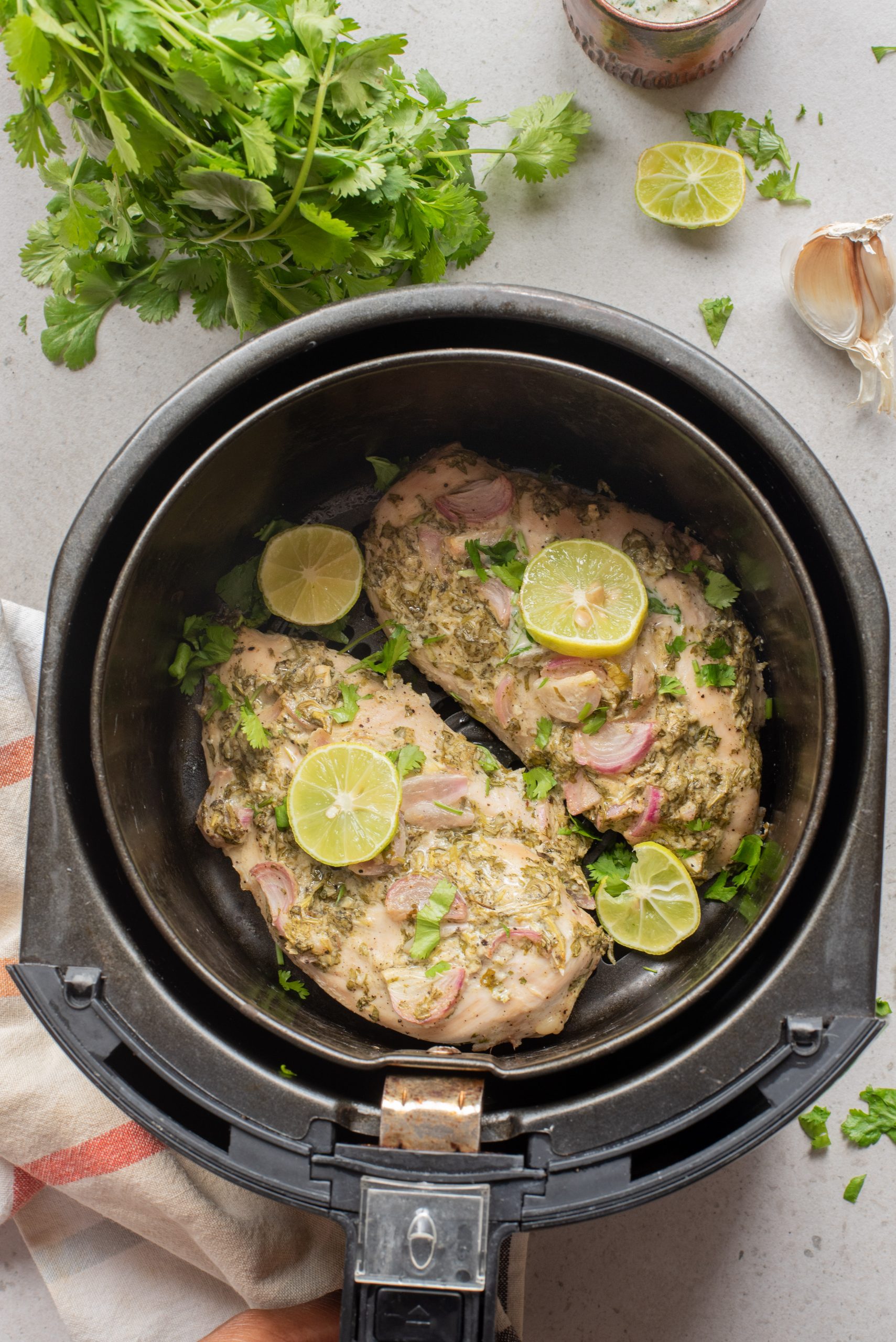 Two seasoned Marinated Yogurt Herb Chicken breasts topped with lime slices and herbs in an air fryer basket, with cilantro, garlic, and a towel nearby on the counter.